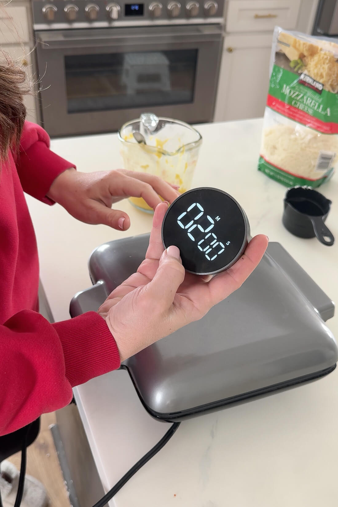 A woman in a red sweater holds a sleek digital kitchen timer set for two minutes, standing beside a closed waffle maker. The countertop has a measuring cup and a bag of shredded cheese.